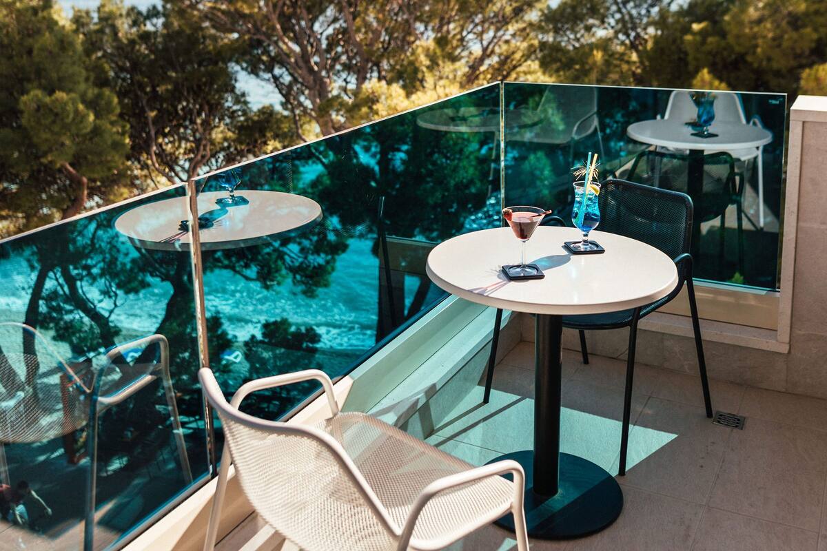 A glass-enclosed balcony with a table and chairs, overlooking a scenic view of trees and the sea.