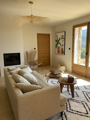 Een woonkamer met een beige bank, een geweven stoel, een ronde houten tafel, een schilderij van planten, en een deur die leidt naar een balkon met uitzicht.