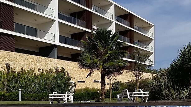 Une piscine à l’eau d’un bleu cristallin, entourée d’un bâtiment moderne doté de nombreux balcons, avec un palmier en premier plan.