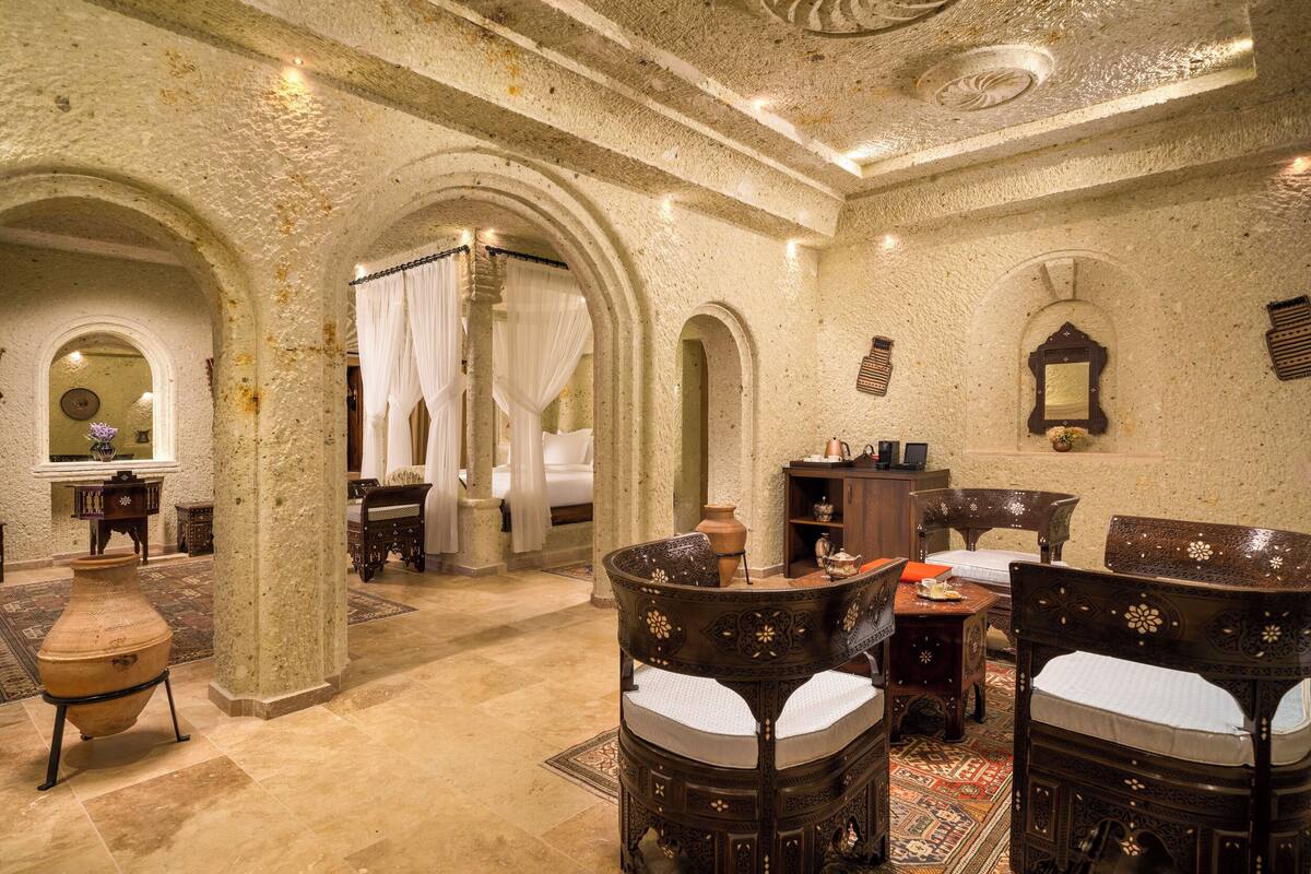 A room with a large arched doorway, a bed with white linens, a wooden chair, a small table with a vase, and a mirror on the wall.