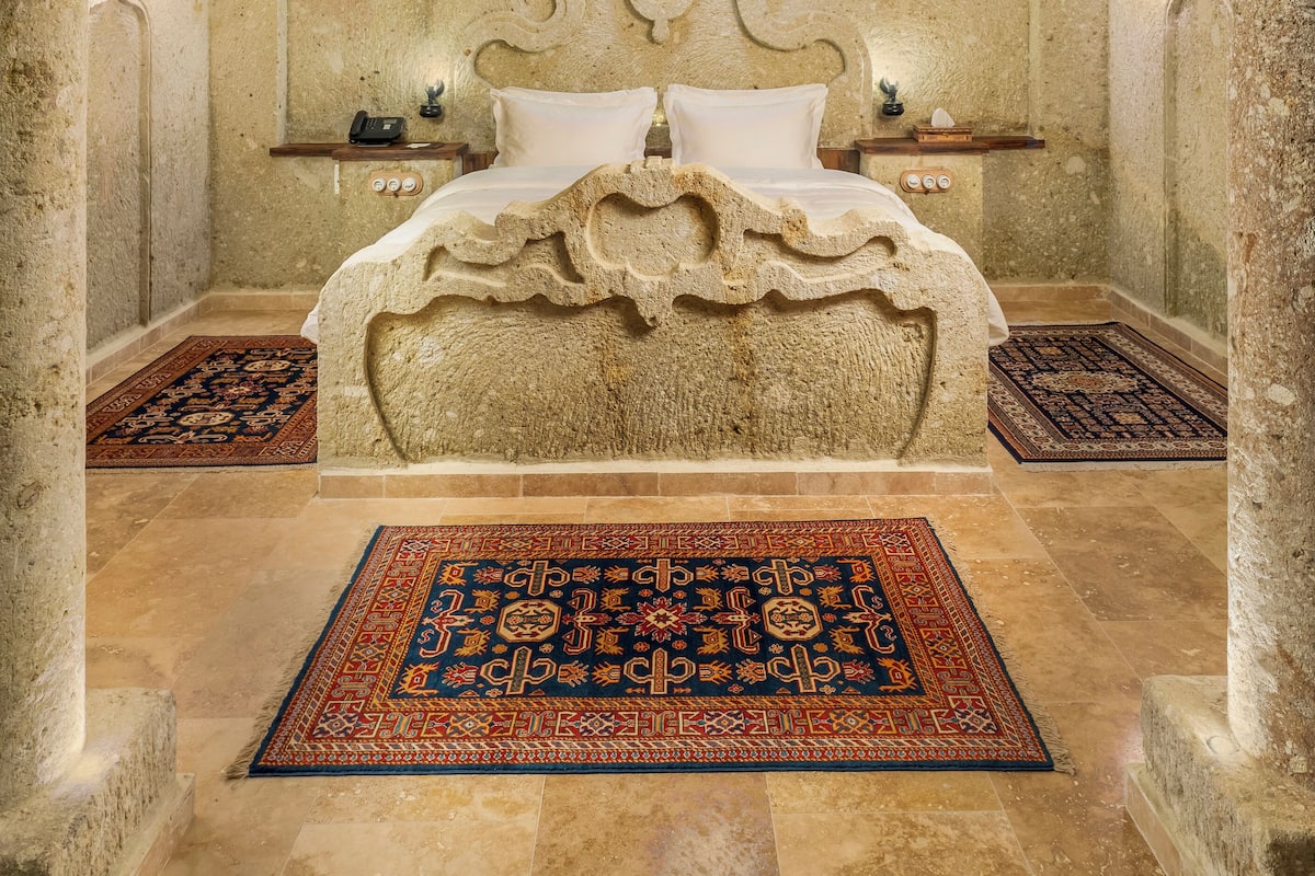 A bedroom with a stone headboard, a bed with white linens, two bedside tables, and two patterned rugs.