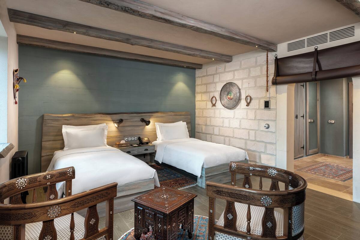 A hotel room with two beds, a wooden headboard, a nightstand, a small table, and a chair. There is a wall-mounted clock and a decorative plate on the wall.