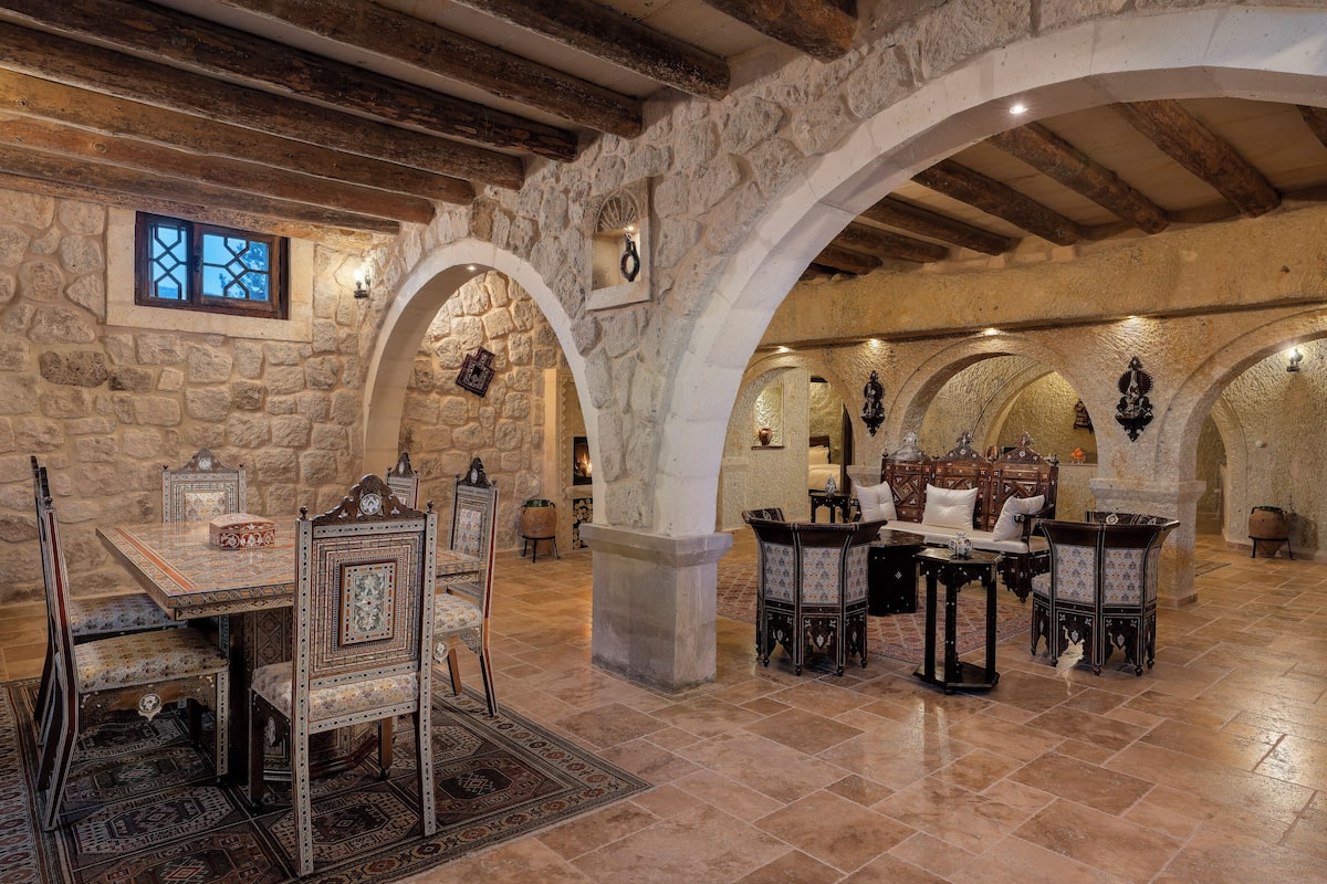 A stone-walled dining area with a high wooden ceiling, rustic furniture, and a patterned rug.