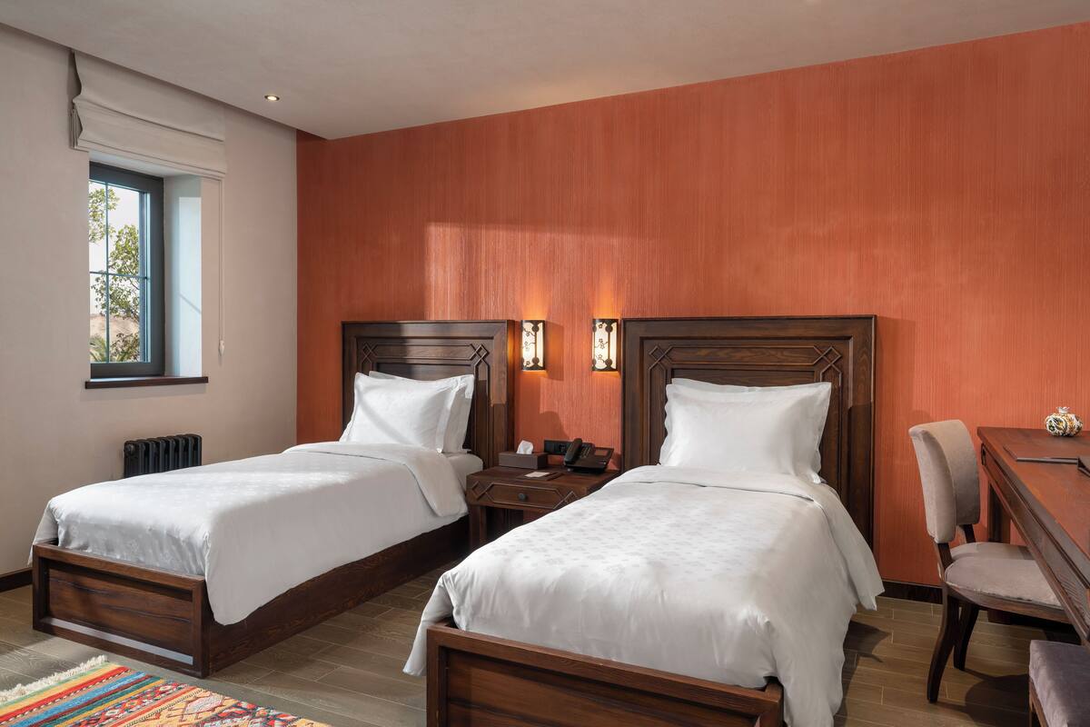 A hotel room with two beds, a desk, a chair, and a window.