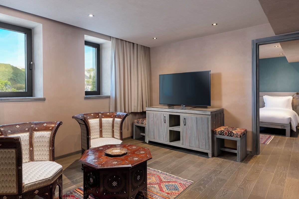 A modern hotel room with a large flat-screen TV, two upholstered chairs, a wooden coffee table, a cabinet, and a bed with a nightstand.