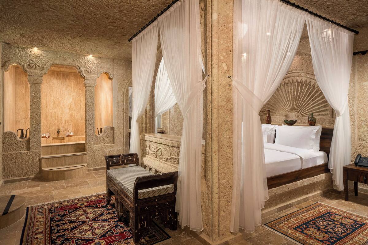 A luxurious bedroom with a four-poster bed, a sitting area with a bench, and a stone wall with a decorative arch.