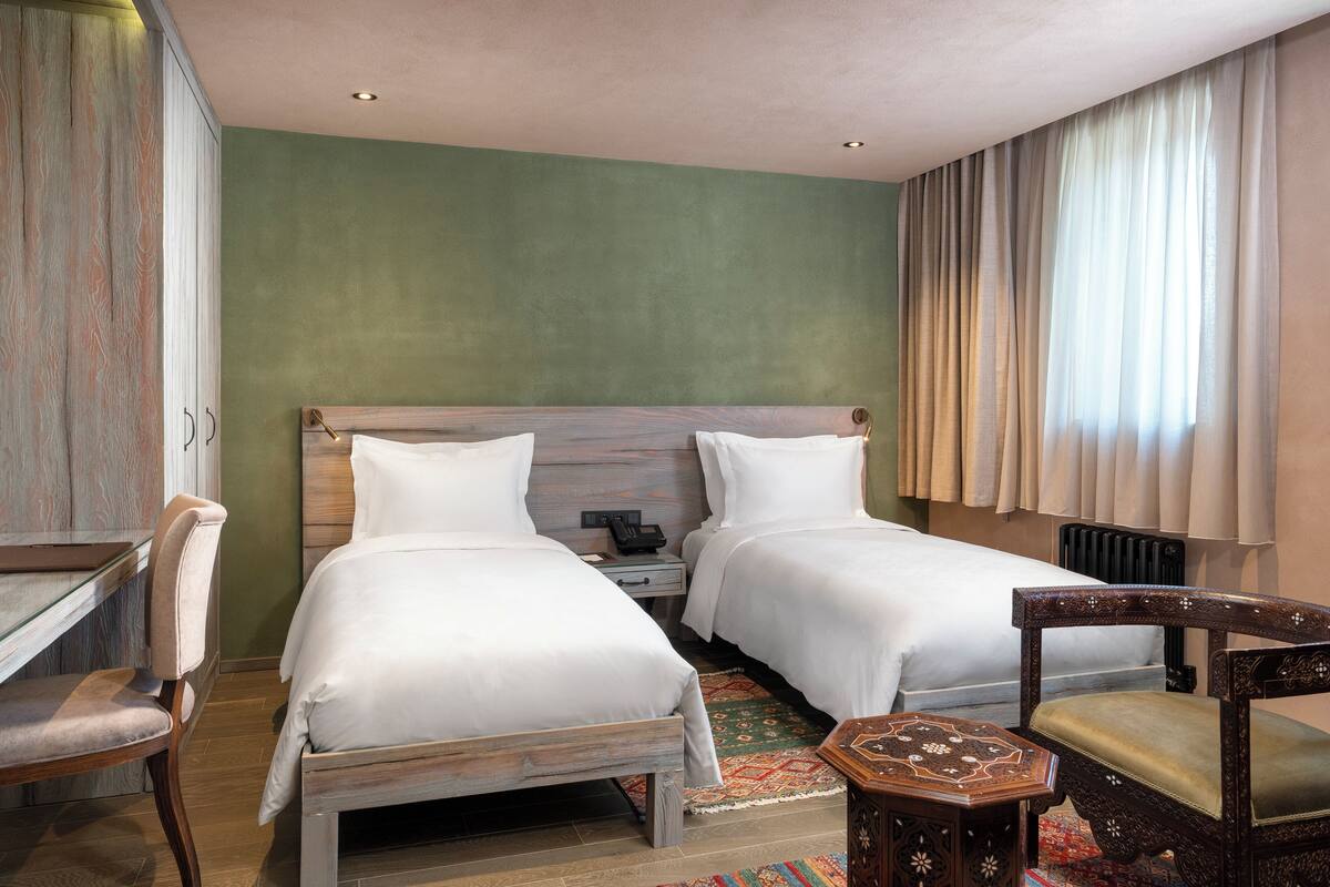 A hotel room with two single beds, a desk, a chair, and a small table.