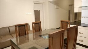 A dining area with a glass table and wooden chairs, a sofa, and a kitchen in the background. - Anmols impex cottage (Ootacamund)
