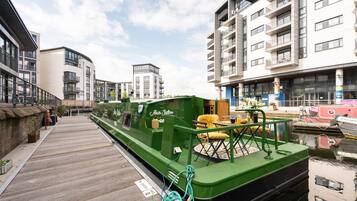 Un barco verde atracado en un muelle con una pasarela de madera, rodeado de edificios modernos y un cielo despejado.
