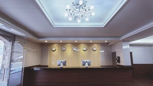 A reception area with a wooden reception desk, a ceiling light fixture, and wall clocks. - Hotel Nueva Europa (Torredembarra)
