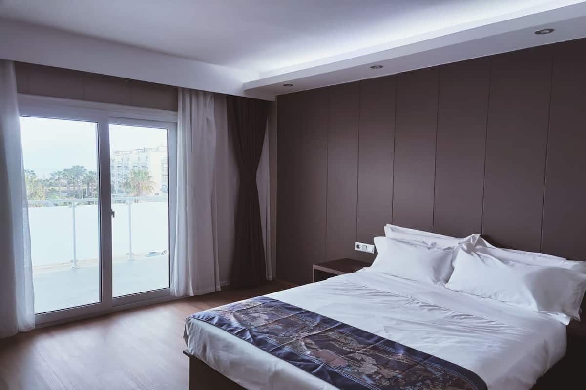 Une chambre d’hôtel moderne avec un grand lit, une petite table de chevet et une vue sur l’extérieur, accessible par une porte-fenêtre coulissante.