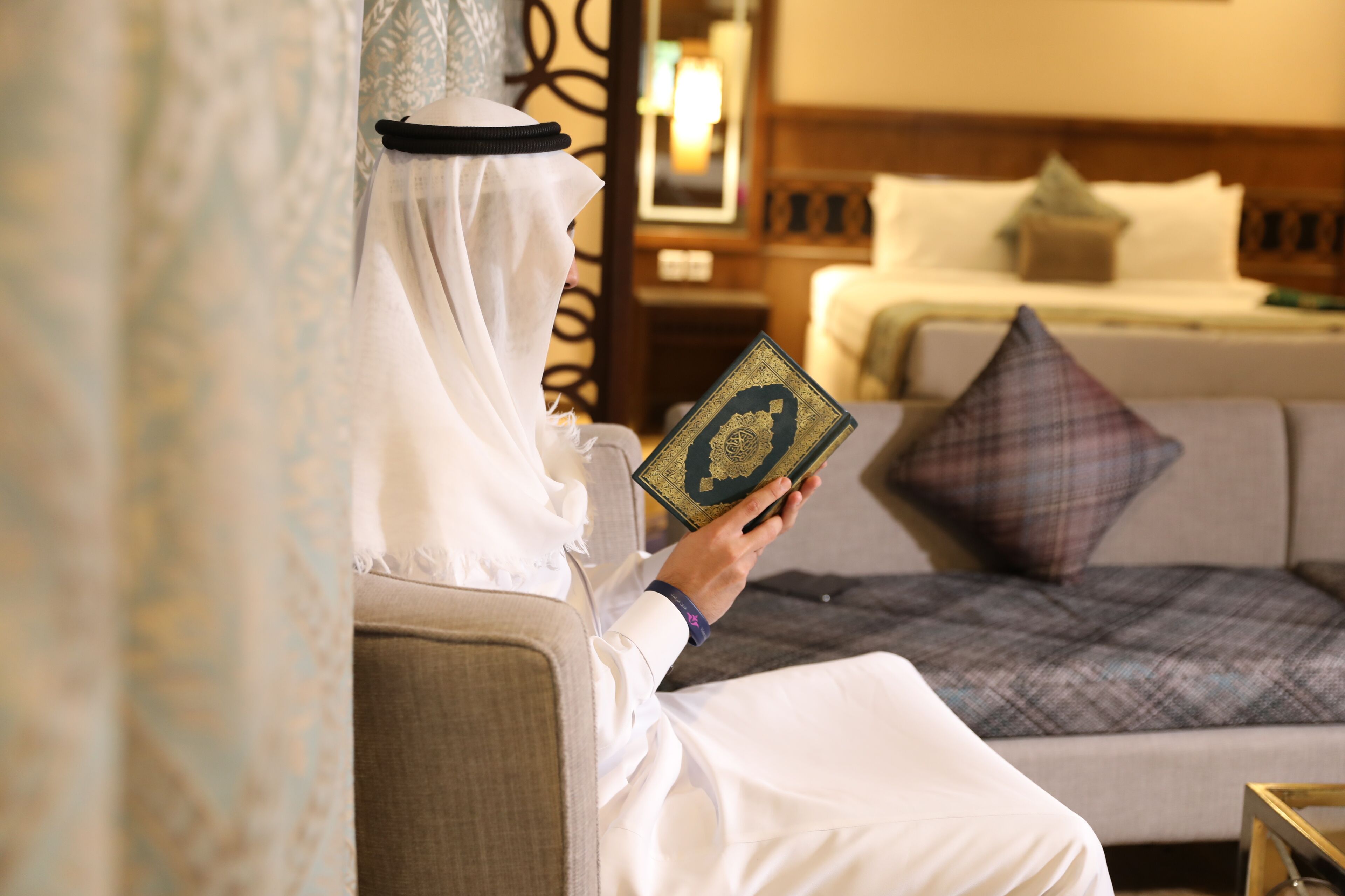 Una persona vestida con traje tradicional leyendo un libro en una habitación de hotel, con cama, almohadas y mesita de noche.