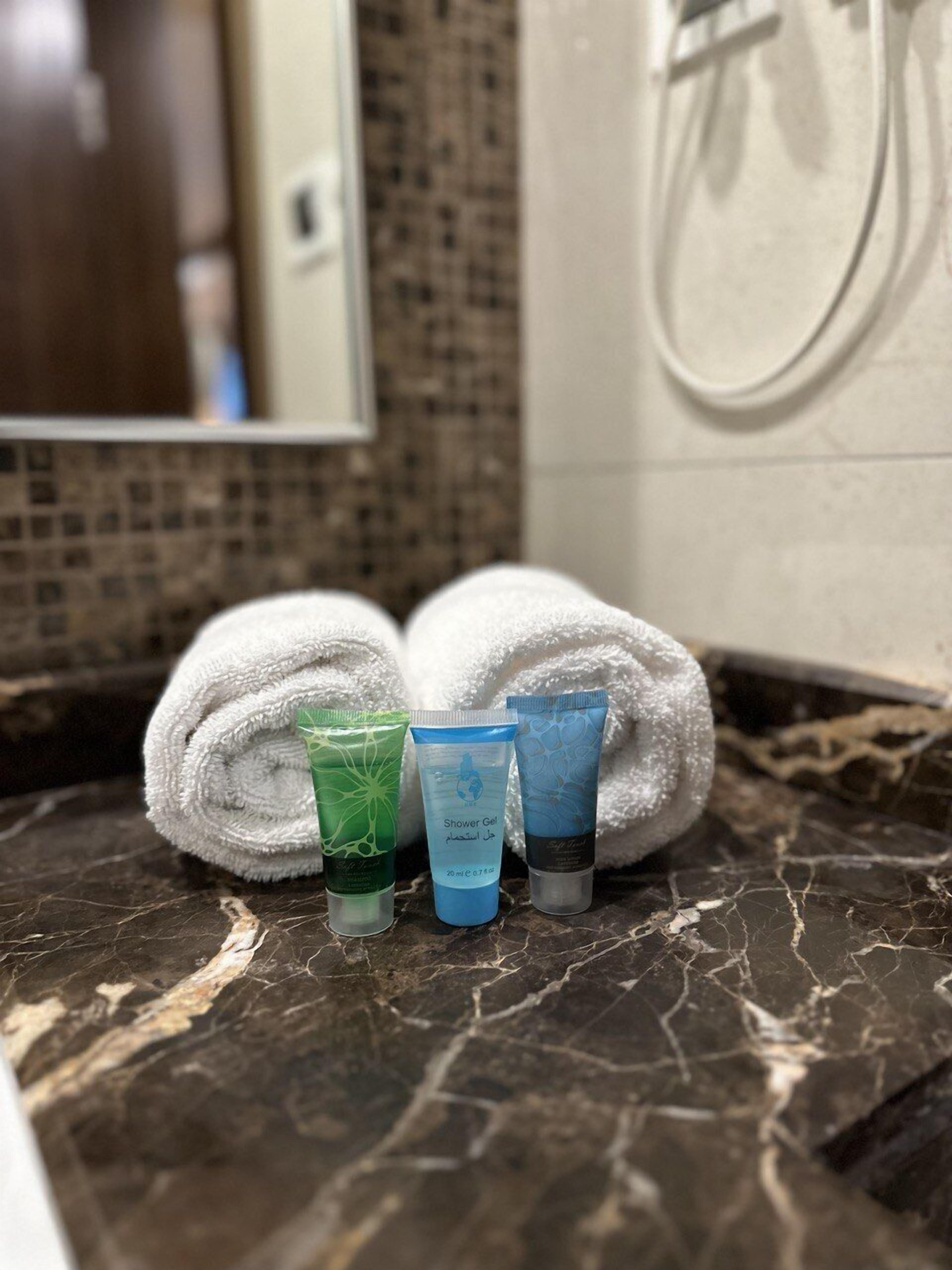 A bathroom with a marble countertop, two rolled white towels, and three toiletries in a green, blue, and grey tube.