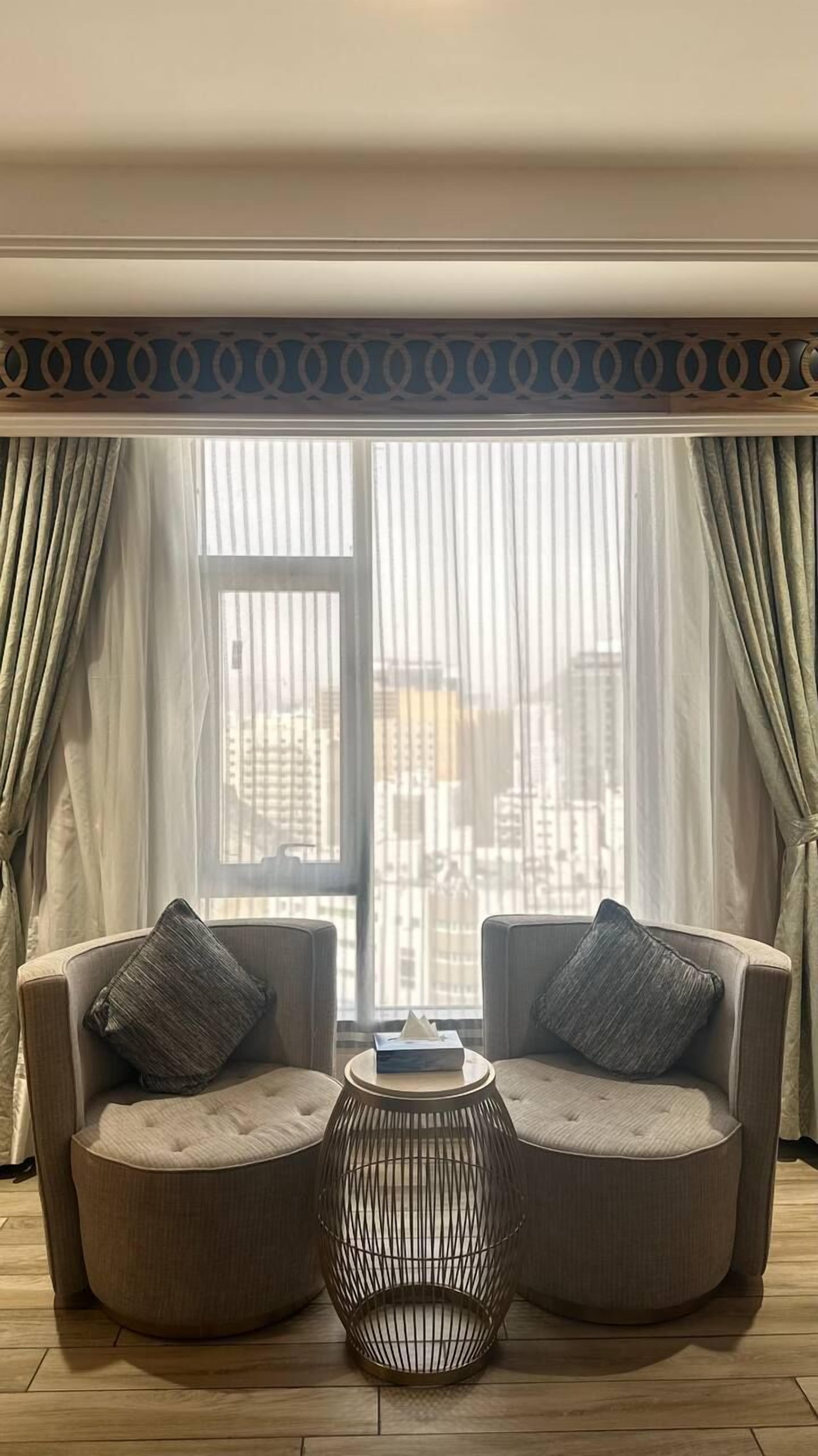Ein Zimmer mit zwei Sesseln, die zu einem großen Fenster blicken, einem kleinen runden Tisch und einer dekorativen Metalllampe.