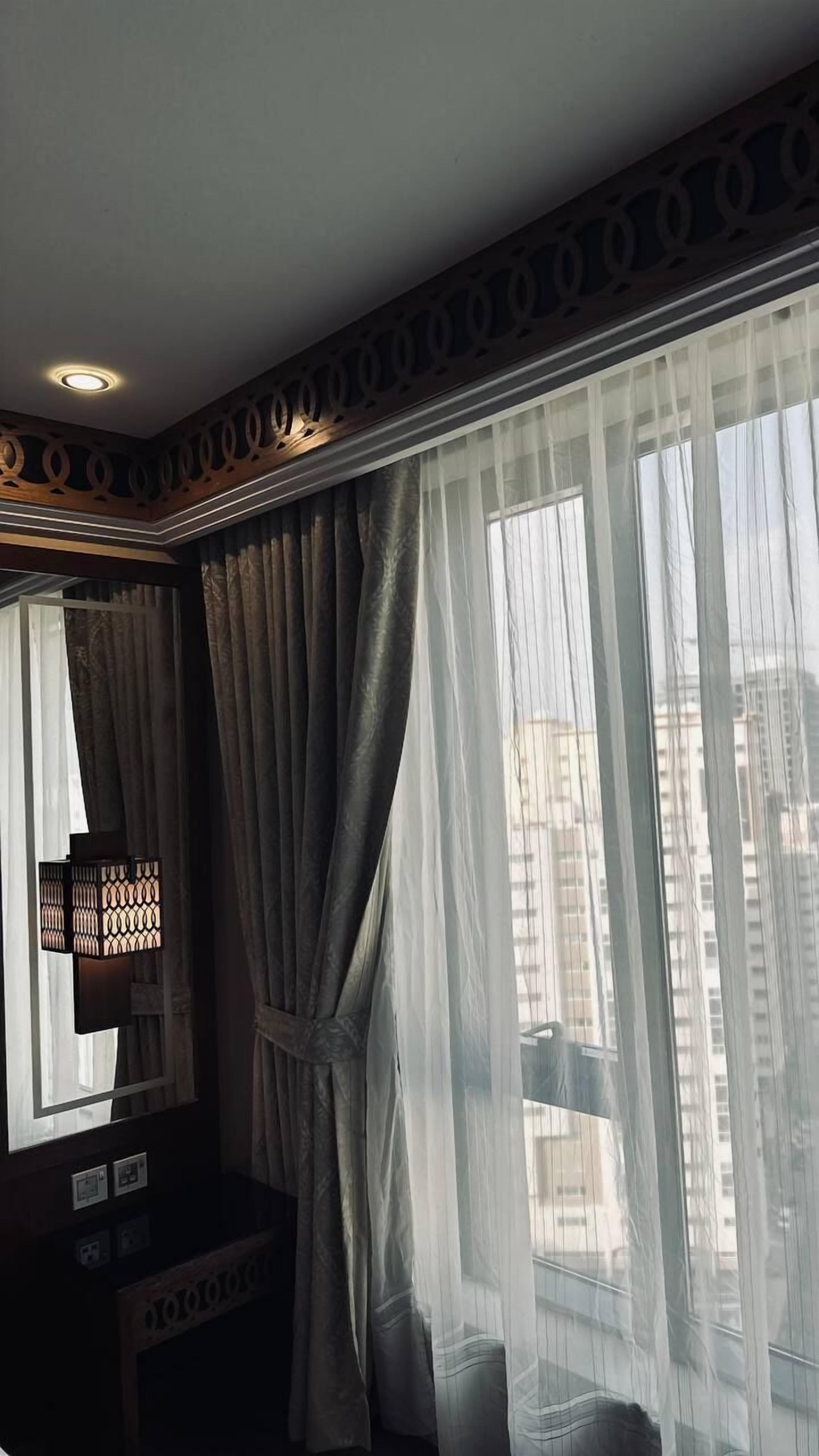 Habitación de hotel con un ventanal grande, cortinas traslúcidas, un espejo y un techo decorativo.
