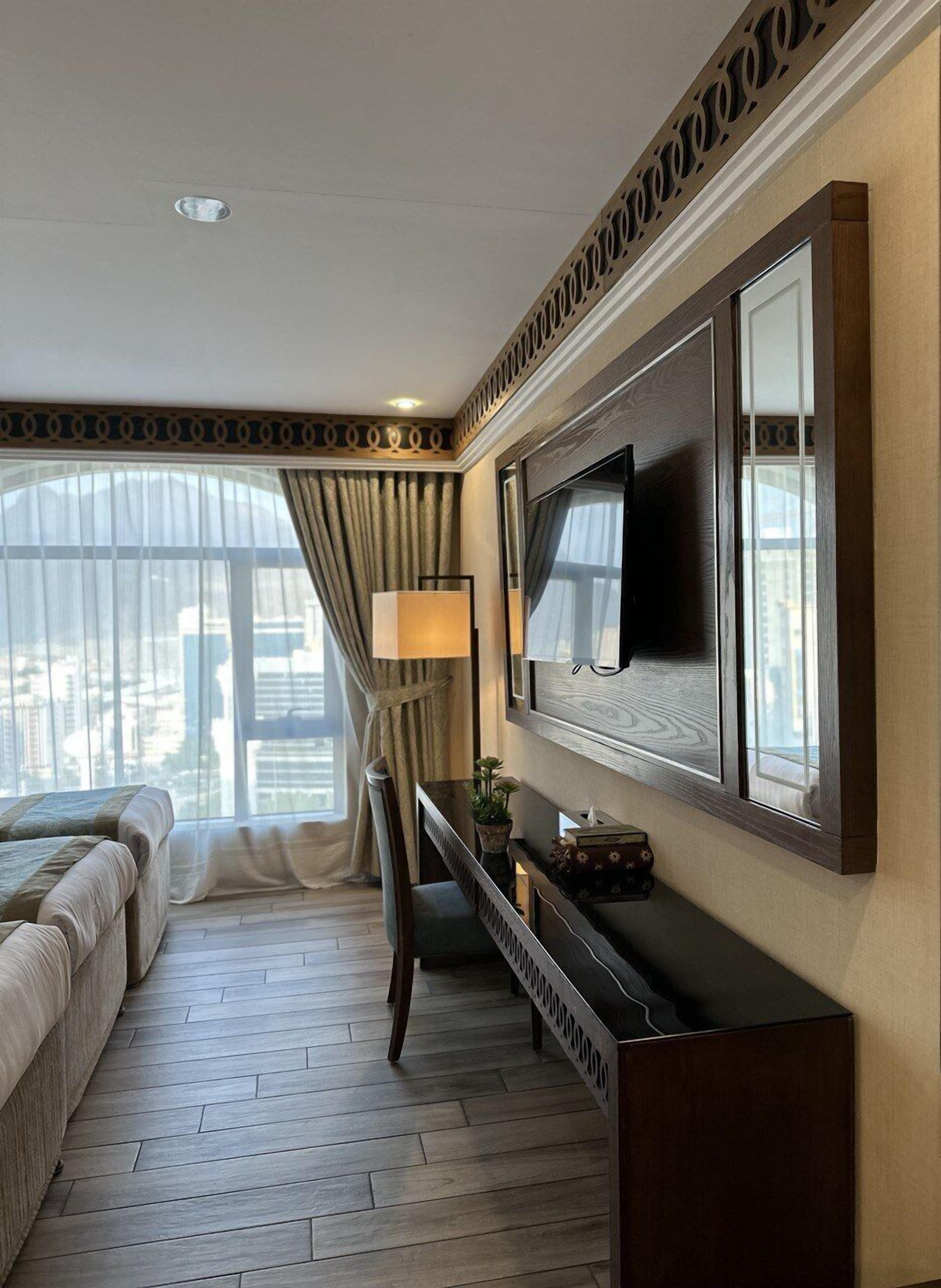 A hotel room with a large bed, a desk with a lamp, a TV mounted on the wall, and a view of the city through large windows.