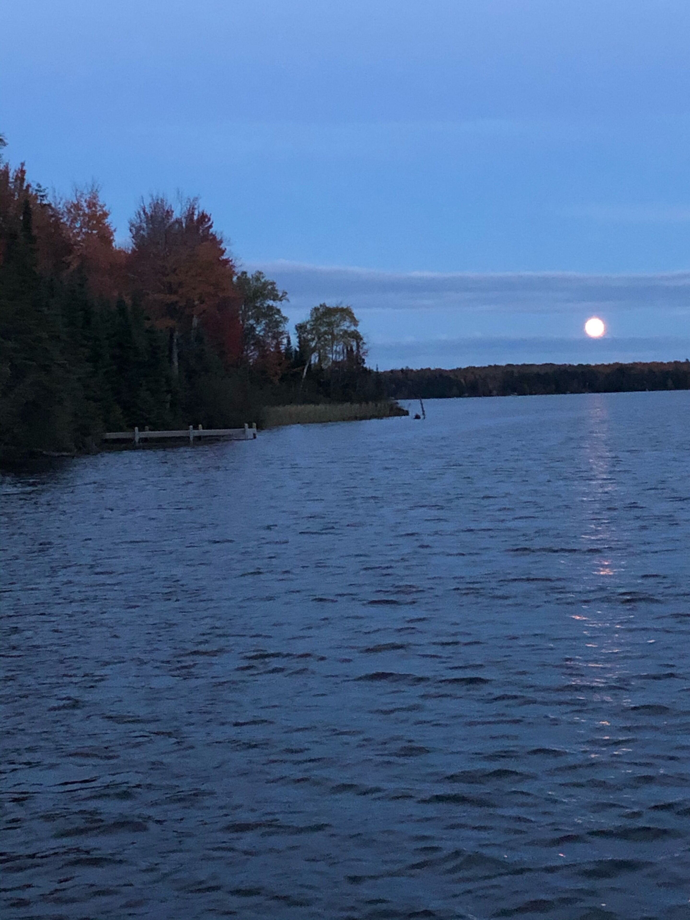 A calm lake with a wooden dock extending into the water, surrounded by trees with autumn foliage.