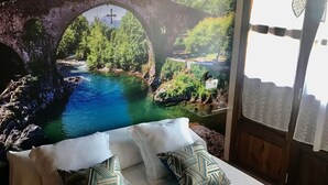 Un llit amb roba de llenceria blanca, coixins estampats, una manta blava i un mural d'un pont sobre un riu.