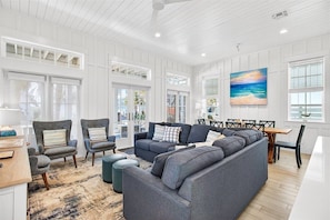 A spacious living room with a large sectional sofa, multiple armchairs, a dining table, and a painting on the wall.