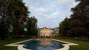 A large, classical building with a symmetrical facade, surrounded by lush greenery and a central swimming pool.