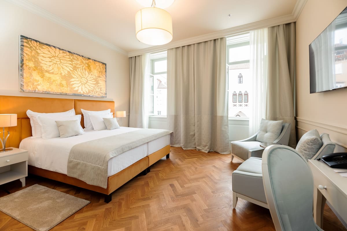 A hotel room with a bed, bedside tables, a desk, a chair, a large window with curtains, and a wall-mounted TV.