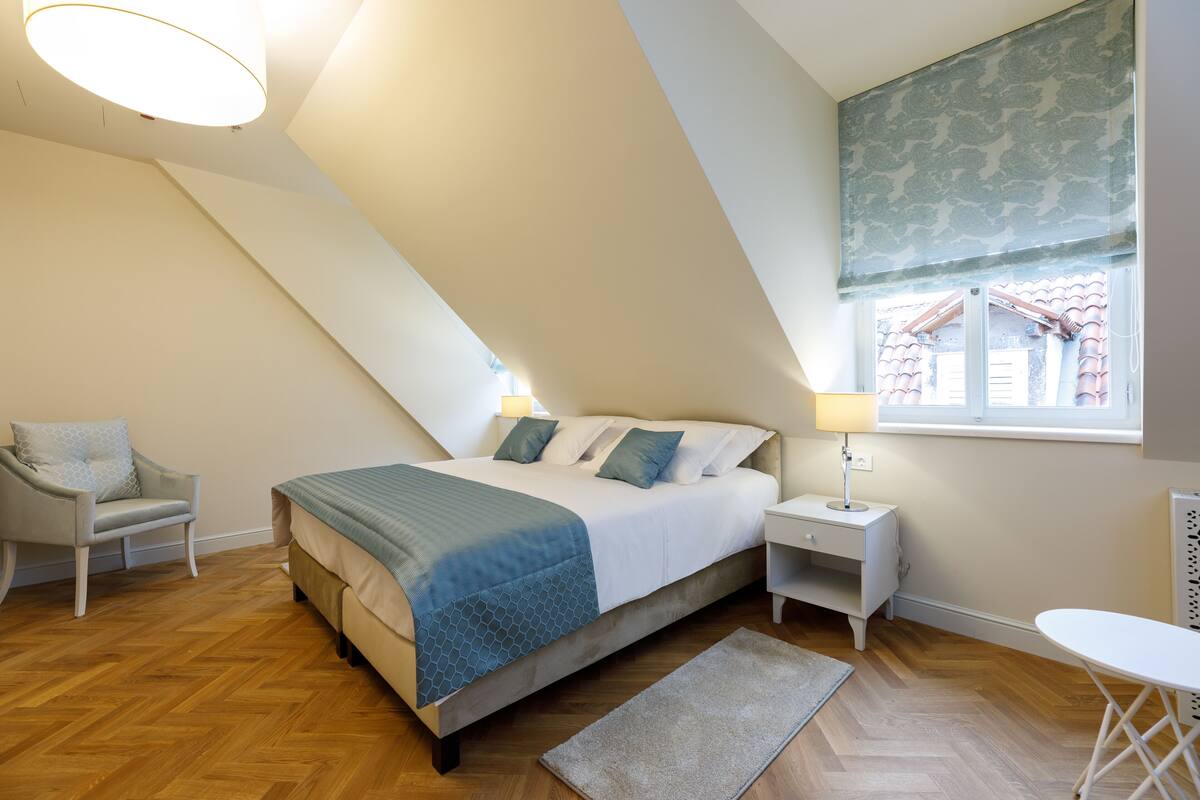 A bedroom with a bed, a chair, a nightstand, a lamp, a window, and a view of buildings outside.