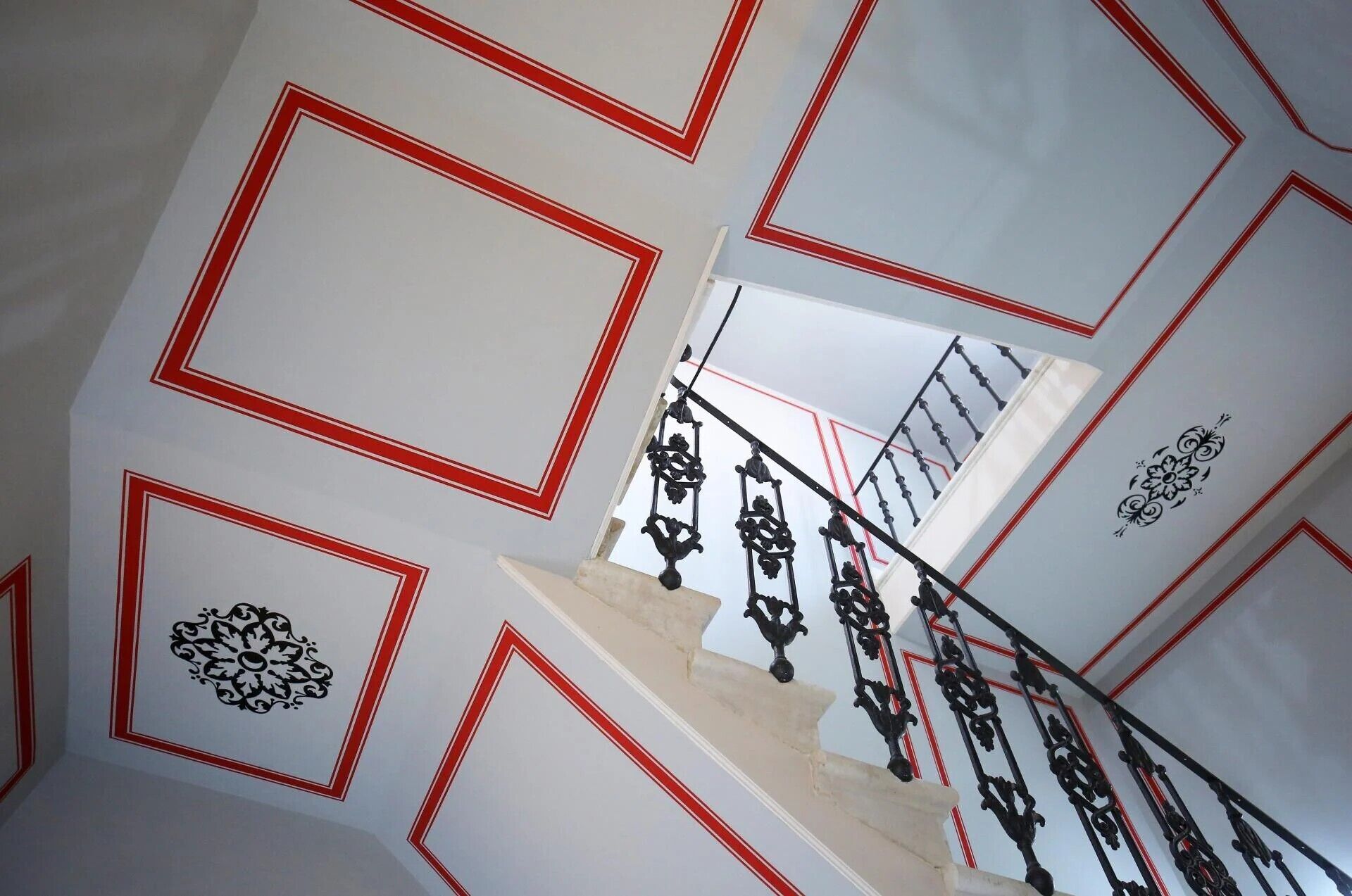 Una escalera con barandales negros ornamentados y paredes blancas con motivos geométricos rojos.