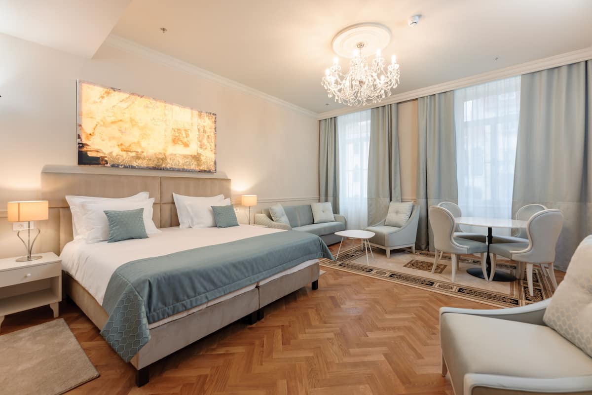 A hotel room with a large bed, a sofa, a dining table with chairs, a chandelier, and a large abstract painting on the wall.