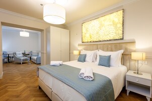 Une chambre d’hôtel avec un grand lit, deux tables de chevet, un bureau avec une chaise et un coin salon avec des fauteuils.