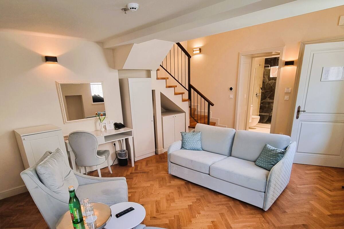 A modern living room with a sofa, armchair, and a small table with a bottle and glasses. A staircase leads to a bathroom.