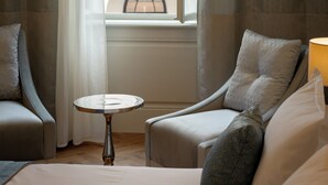 Habitación de hotel con cama, dos sillones, una mesita y ventana que ofrece vistas a un edificio con ventanas de arco y un balcón.