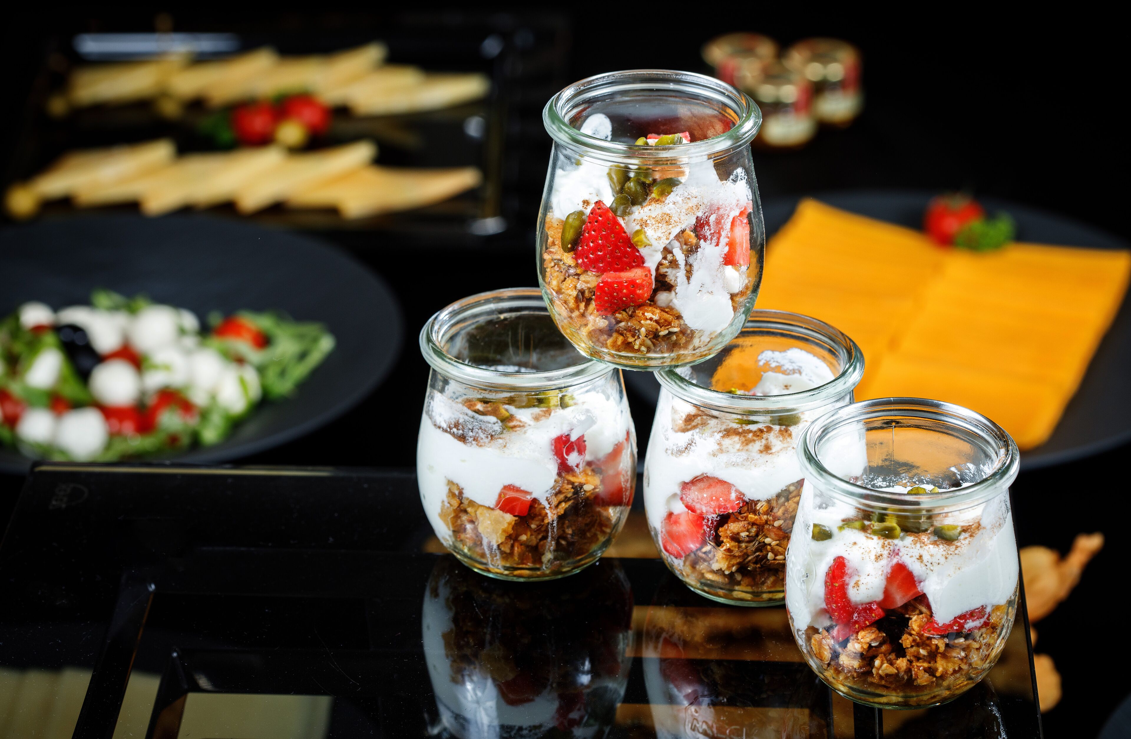 Four glass jars containing layered granola with strawberries and yogurt, a plate of salad, and a piece of cheese on a reflective surface.