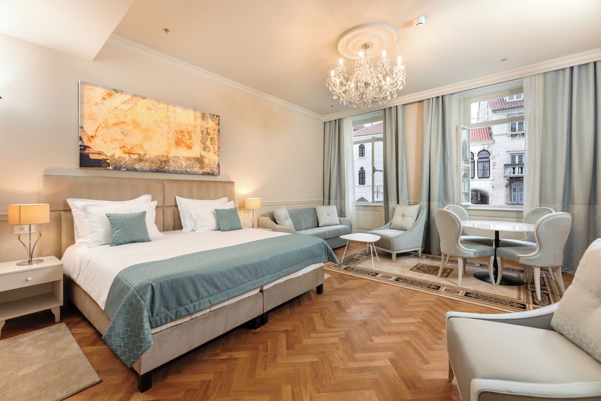 A hotel room with a bed, a sofa, a dining table with chairs, and a view of buildings outside.