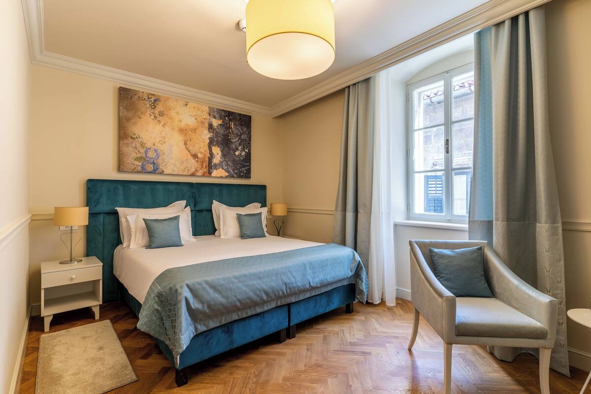 A neatly arranged bedroom with a large bed, a chair, a nightstand, and a window with curtains.