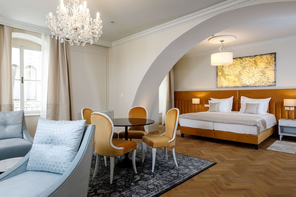A hotel room with a bed, a sofa, a dining table with chairs, a chandelier, and a large painting on the wall.