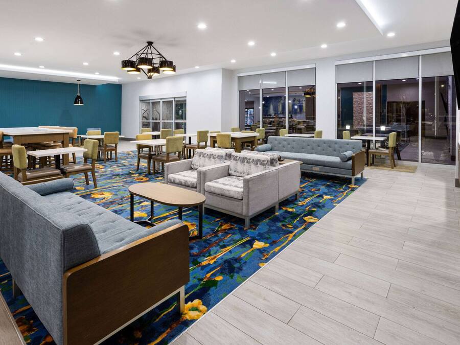 Un hall d’hôtel moderne avec un mur d’accent bleu, divers types de sièges, notamment des canapés et des fauteuils, une moquette à motifs et un grand écran de télévision fixé au mur.
