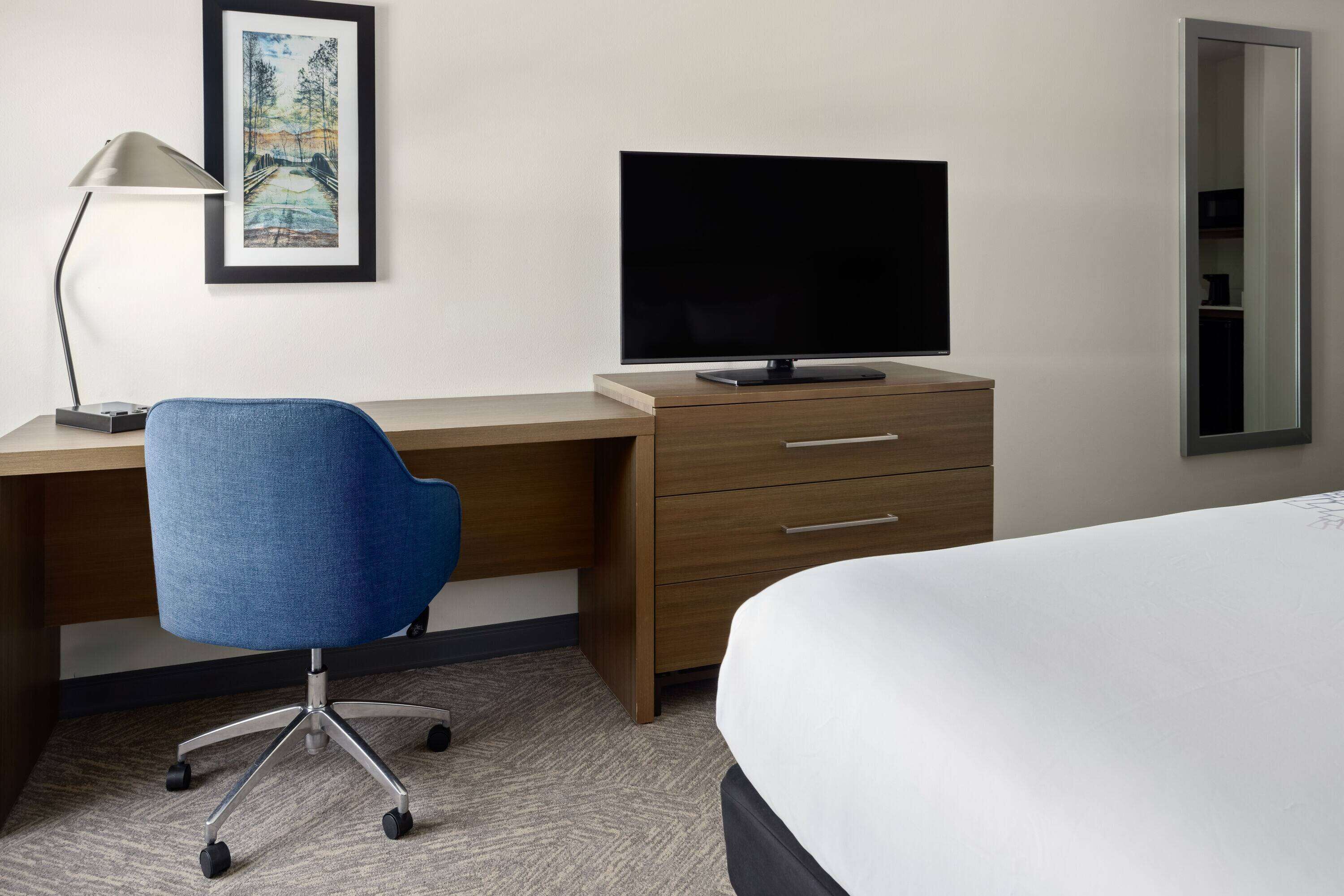Habitación de hotel con escritorio, silla, televisión, cama y un cuadro en la pared.