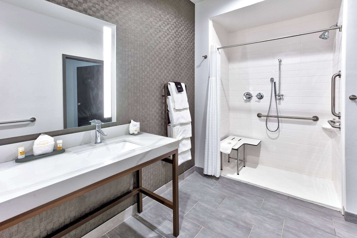 A modern bathroom with a large mirror, a sink with toiletries, a shower with grab bars, and a towel rack.