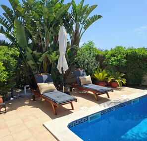 Un área junto a la piscina con dos sillones de descanso, una piscina pequeña y abundante vegetación.