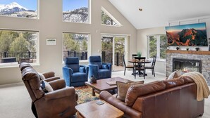 A spacious living room with a fireplace, leather sofas, a coffee table, and a TV. Large windows offer mountain views. - Secluded Creekside Home | Incredible Views | A/C | Private Deck | STR2025-036 (Ridgway)