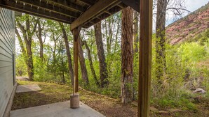 A covered outdoor area with a concrete floor, surrounded by trees and a hillside in the background. - Secluded Creekside Home | Incredible Views | A/C | Private Deck | STR2025-036 (Ridgway)