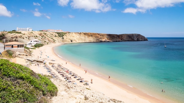 Une plage côtière avec un rivage sablonneux, des eaux turquoise et limpides, et une falaise rocheuse.