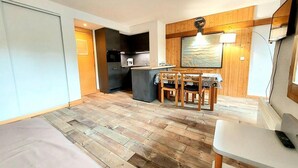 A compact room with a bed, a dining table with chairs, a kitchen area, and a TV mounted on the wall.