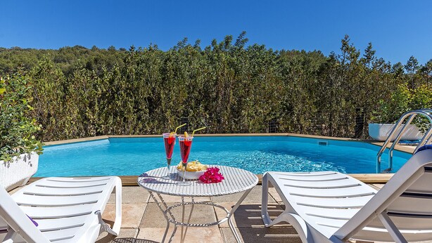 Zona de piscina con vista despejada, rodeada de exuberante vegetación. Hay sillas de descanso y una mesa con bebidas y un arreglo floral.