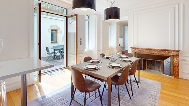 A dining area with a table set for six, two pendant lights, a fireplace, and a view through a glass door to an outdoor seating area.