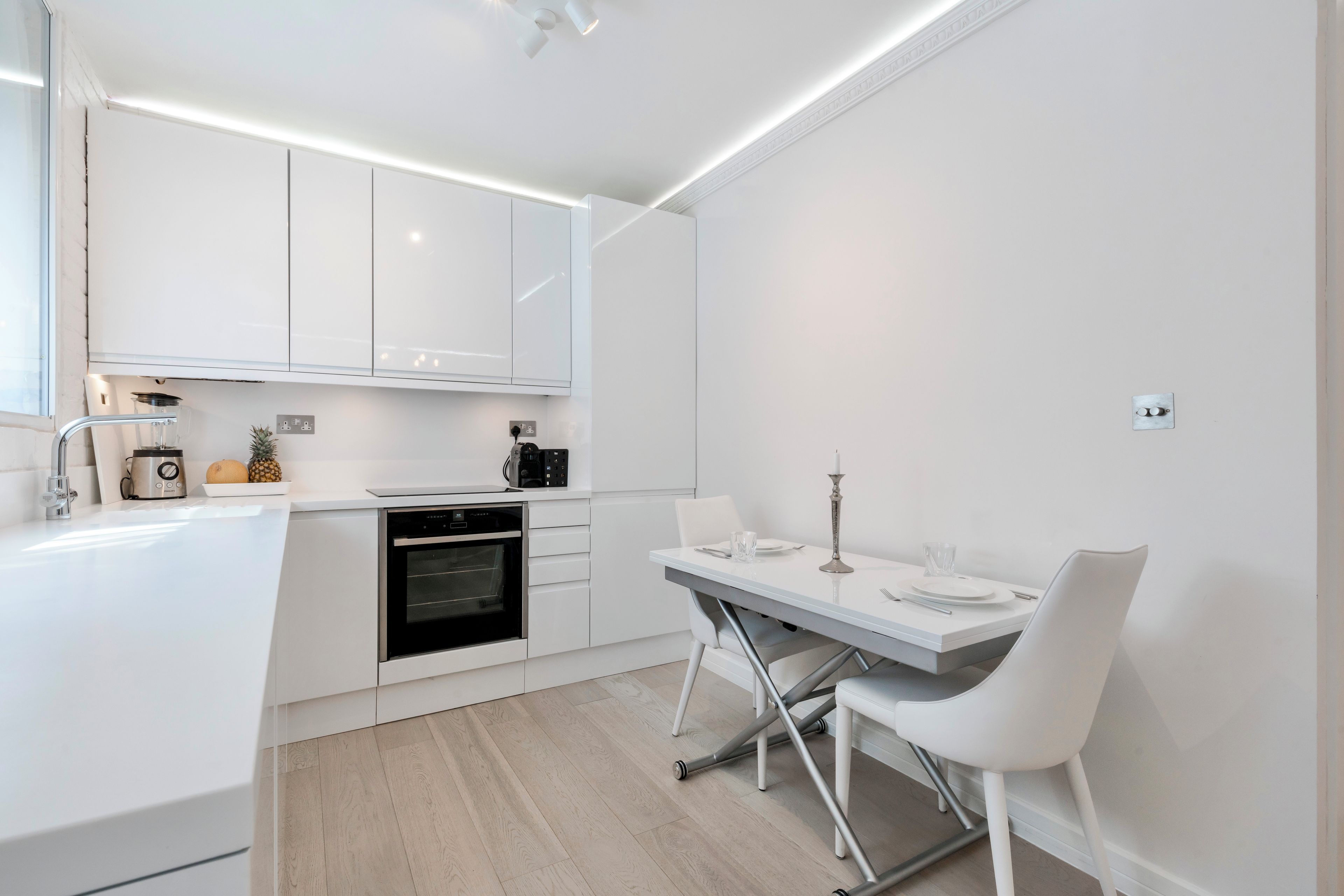 Een moderne keuken met witte kasten, een ingebouwde oven en een eethoek met een witte tafel en stoelen.