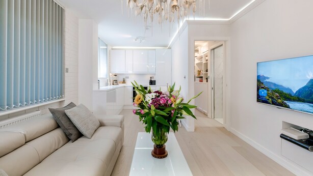 Een moderne woonkamer met een bank, een bijzettafeltje met een bloevas, een televisie aan de muur en een keuken op de achtergrond.