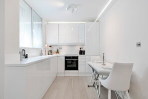 Una cocina moderna con muebles blancos, horno empotrado y comedor con mesa y silla blancos.
