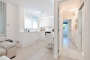 Una cocina moderna con armarios blancos, un comedor con una silla blanca y un pasillo que lleva a un dormitorio.
