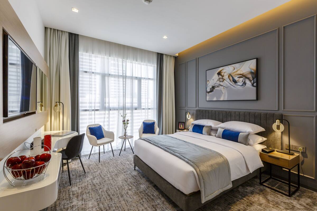 A modern hotel room with a large bed, a desk with a bowl of apples, a chair, a small table with a vase, and a large window with curtains.